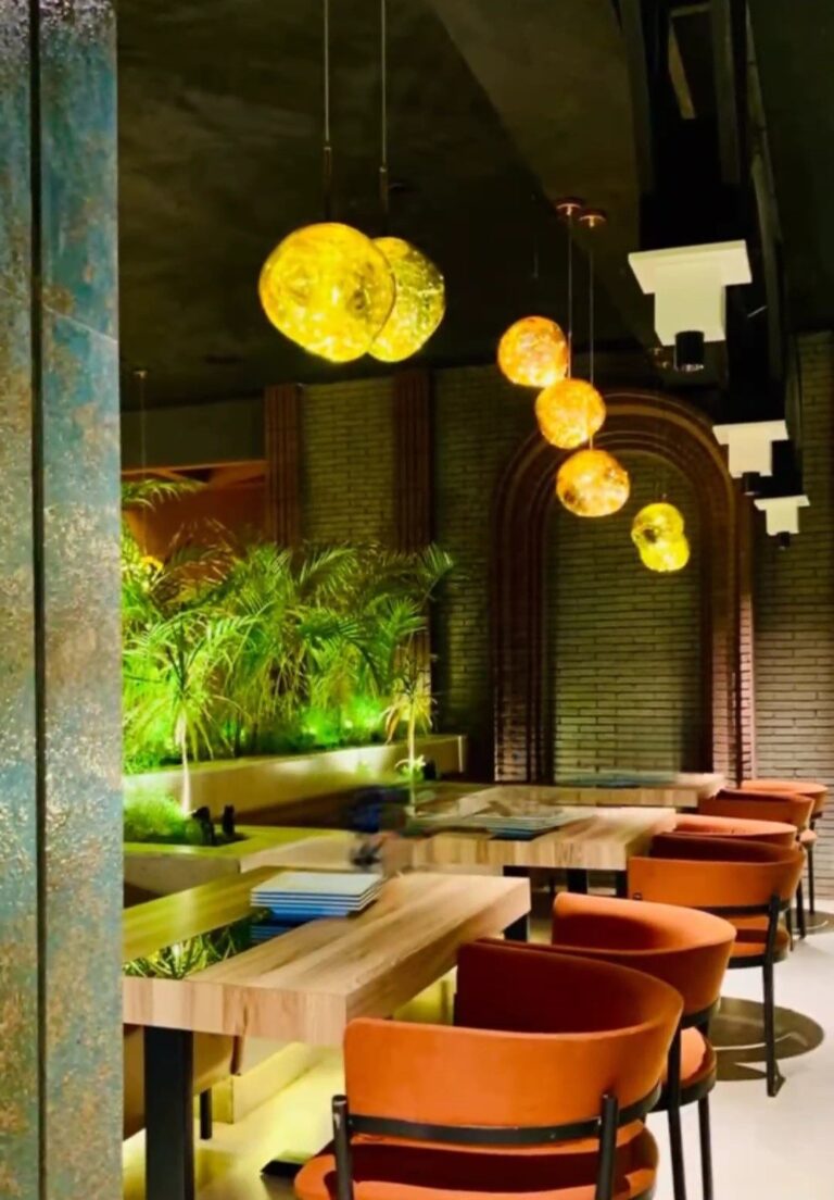 A restaurant dining area with warm wood tables, rust-colored chairs, and spherical pendant lighting beneath a brick archway. Designed by ZM Architects.