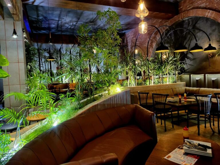 A garden-themed restaurant interior with dense overhead greenery, brick archways, and leather booth seating, designed by ZM Architects.